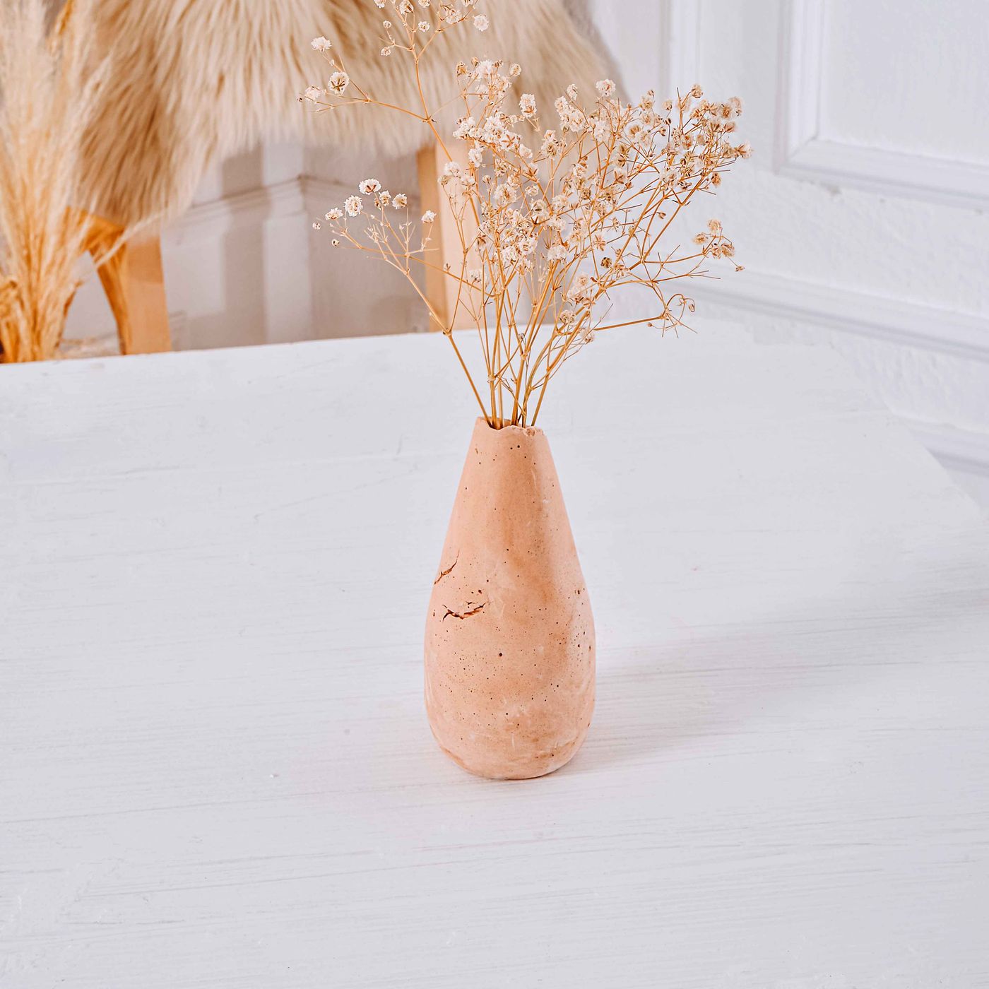 A clay vase photographed in a minimalist setting on a white table, holding a few dried flowers.