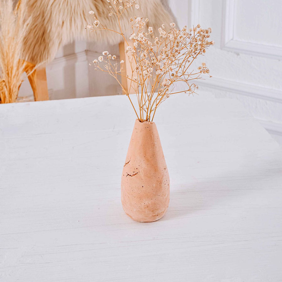 A clay vase photographed in a minimalist setting on a white table, holding a few dried flowers.