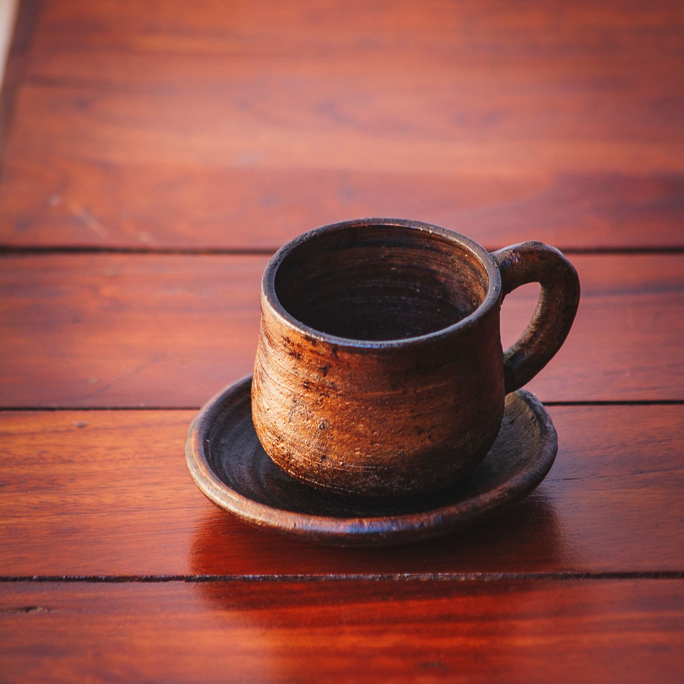 A rustic clay cup with its saucer, photographed together on a wooden table in traditional pottery style.
