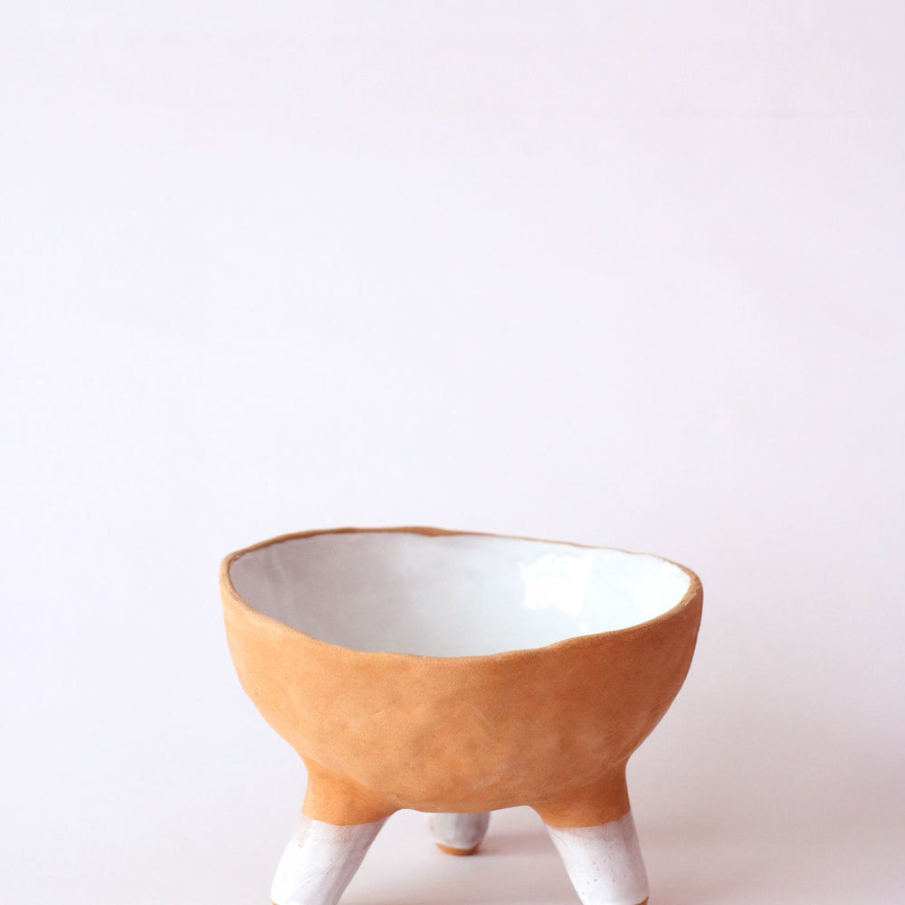 A minimalist ceramic bowl raised on a three legged tripod design, shown on a clean white backdrop.