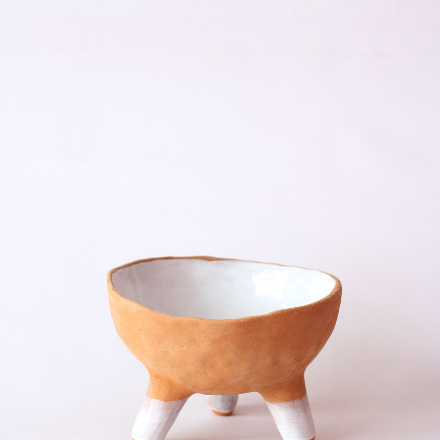 A minimalist ceramic bowl raised on a three legged tripod design, shown on a clean white backdrop.