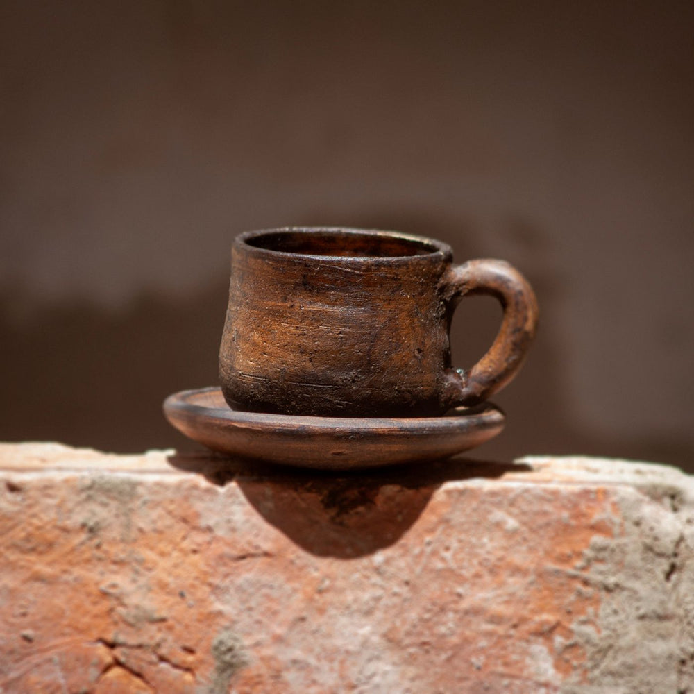 A classic clay coffee cup with its matching saucer, shown against a warm earthy backdrop.