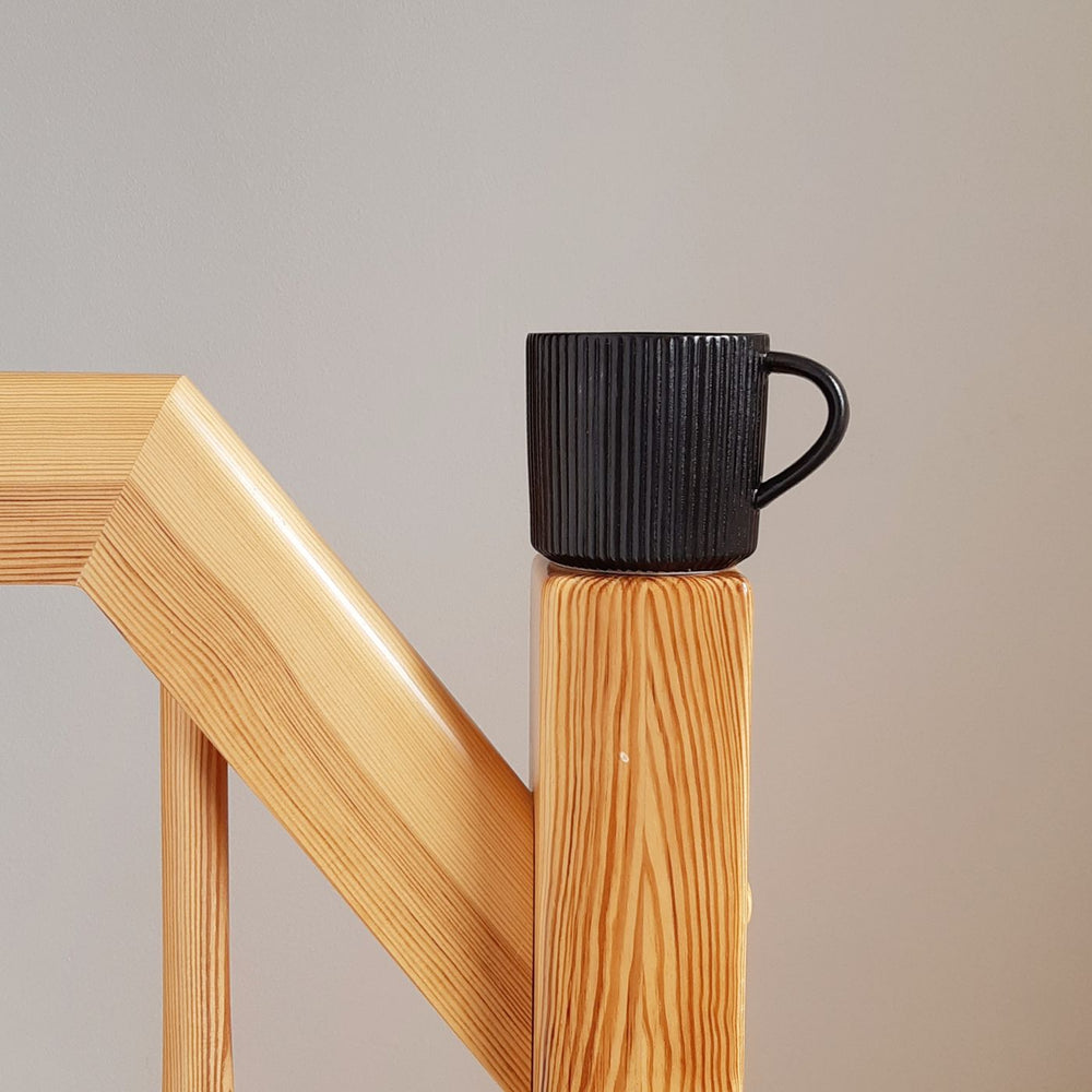 A black ceramic mug with a minimalist shape and earthy tones, shown standing on a wooden railing.