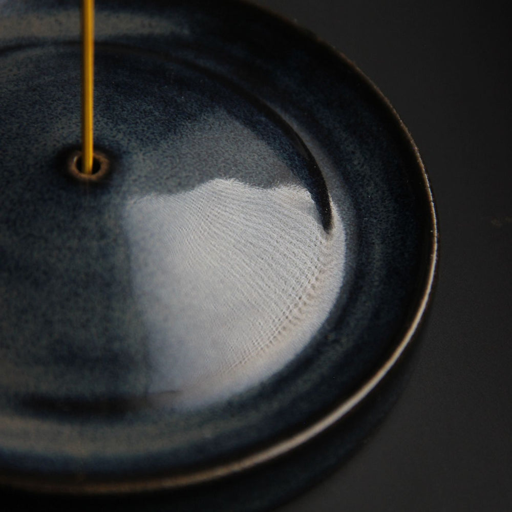 A ceramic incense holder shown close up releasing smoke in a dark minimalist setting.