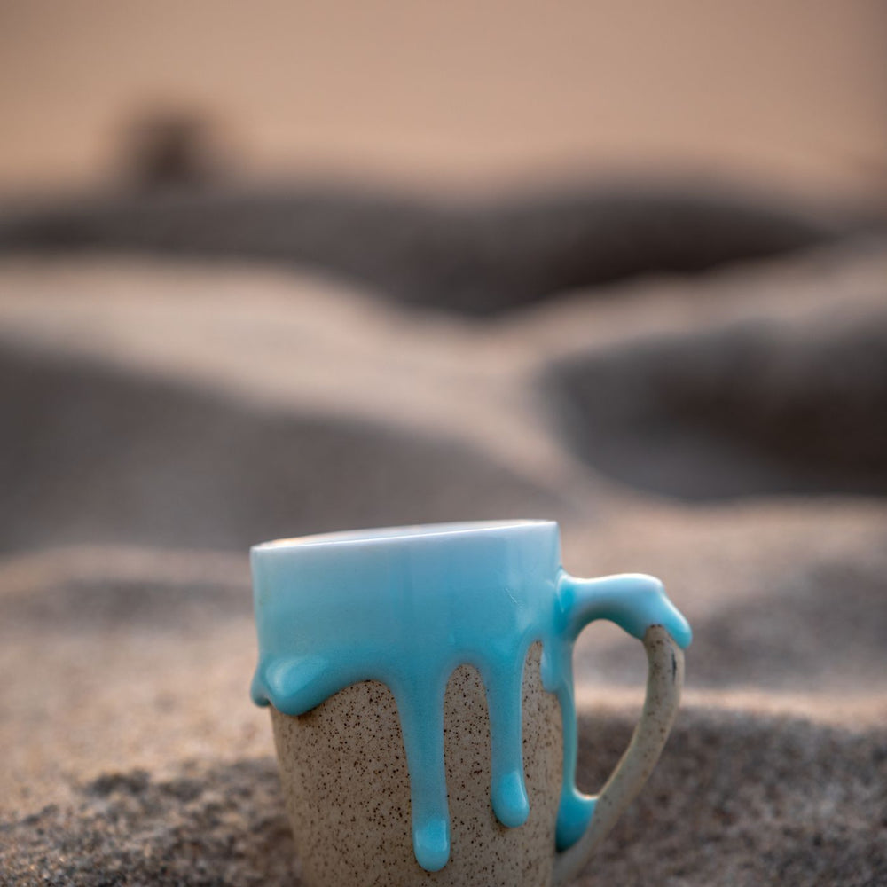 A ceramic mug in a blue glaze, photographed standing in the sand at sunset.