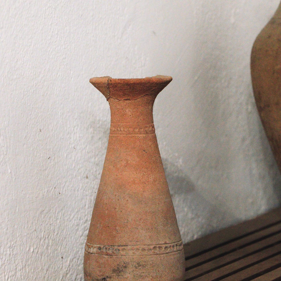 A clay vase in an ancient style, carrying intricate worked design and shown on a wooden shelf.