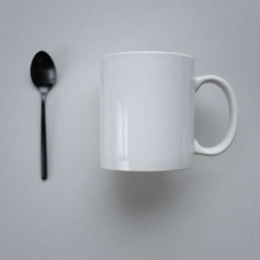 A white mug photographed on a neutral grey background with a black spoon laid beside it.