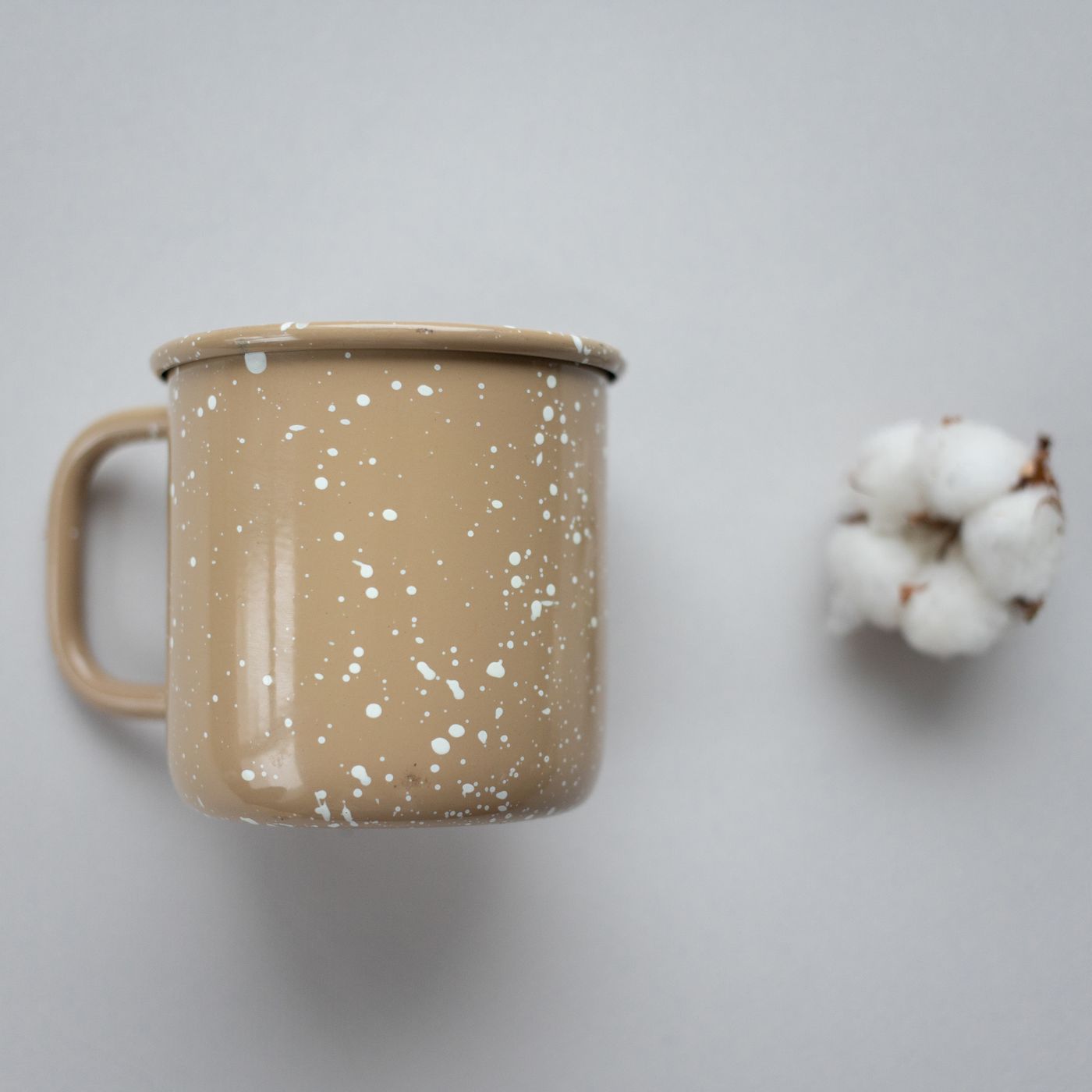 A speckled ceramic mug photographed on a neutral surface beside a stem of cotton blossom.