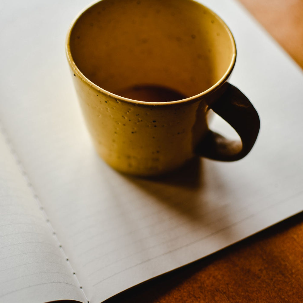 A small brown ceramic cup, shown empty and resting on an open notebook on a wooden desk.