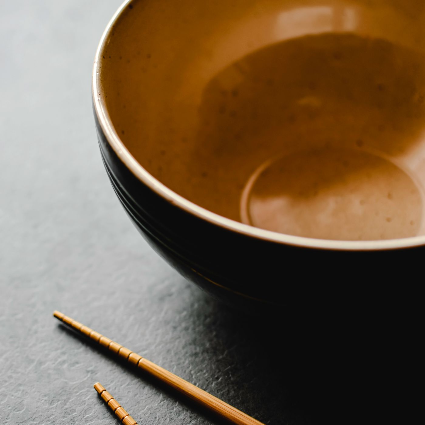 A warm toned ceramic bowl photographed close up on a dark surface with wooden chopsticks resting across it.