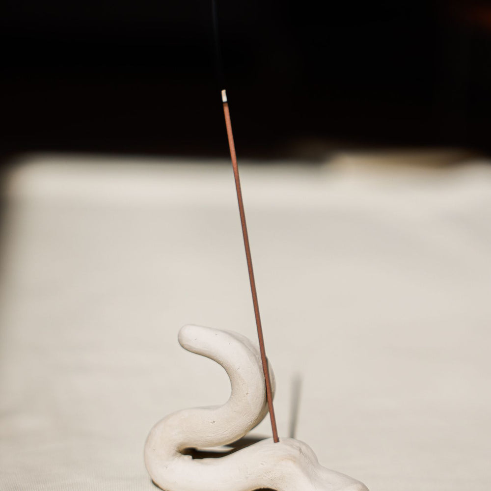 A minimalist ceramic incense holder, photographed with a stick burning in it against a soft background.