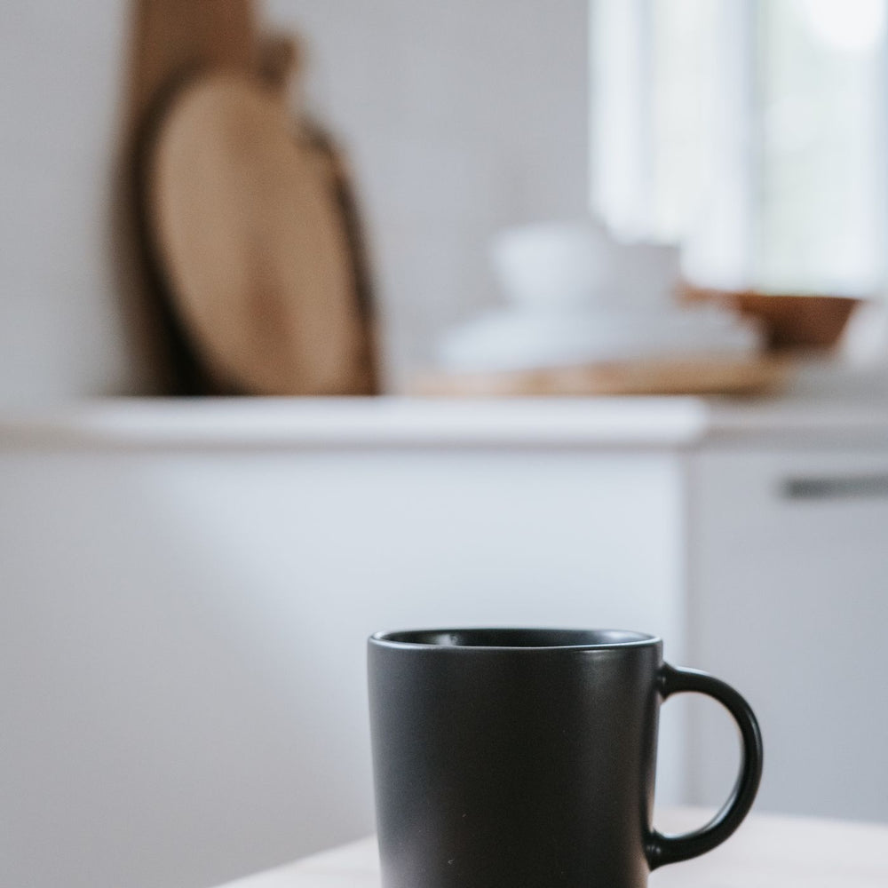 A black ceramic mug photographed on a wooden table in a bright kitchen.